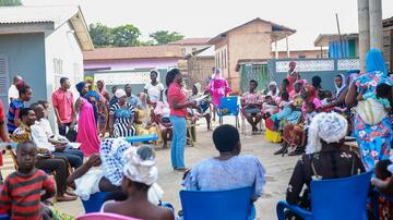 A female health care worker in a red KCCR T-shirt stands in the centre of a circle of around 50 standing and sitting women, men and children in a square surrounded by a few houses.