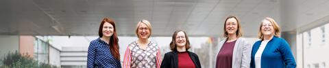 Fünf Frauen stehen lächelnd in einer Reihe vor einem modernen Bürogebäude. Sie tragen unterschiedliche Business-Outfits und blicken selbstbewusst in die Kamera.