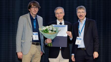 Jan Rupp and Dirk Busch flank award winner Christoph Lange at the award ceremony. All three stand on a stage against a dark background. Christoph Lange holds a bouquet of flowers and a certificate in his hands.