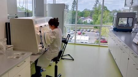 View into a laboratory of the MiFactR project: On the left of the picture, a woman in a white lab coat is sitting at the anaerobic workstation. In the background there is a large window front with a view over a parking lot.
