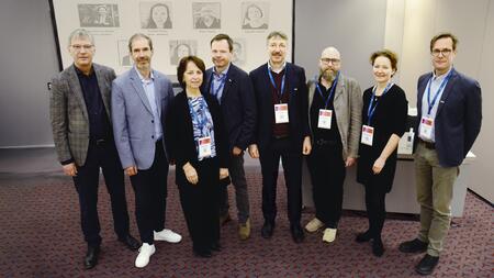 A group of eight people wearing name badges and blue conference lanyards pose for a photo in a conference room. In the background, a screen displays portraits and names of additional people.