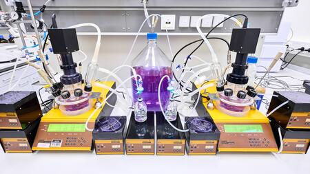 Labor equipment on a workbench with two round glass vessels, many connected tubes, a large bottle with purple liquid in the center, and several electronic control units underneath.