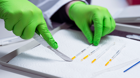 A person wearing green protective laboratory gloves is working on a sterile surface. She is holding a scalpel in one hand and arranging several labeled capillary tubes with yellow caps on a tray with white paper in the other. The scene probably takes place in a laboratory in the context of liquid biobanking or similar sample processing.