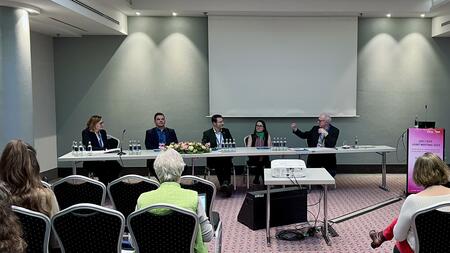 Das Bild zeigt eine Podiumsdiskussion bei einer Konferenz. Fünf Personen sitzen an einem langen Tisch, während eine Person spricht und gestikuliert. Im Vordergrund sitzen einige Zuhörerinnen und Zuhörer. Rechts steht ein Rednerpult mit der Aufschrift „DGI / DZIF Joint Meeting 2025“. Der Raum hat eine schlichte Einrichtung mit einem Projektor, einem großen Bildschirm und Wasserkaraffen auf dem Tisch.