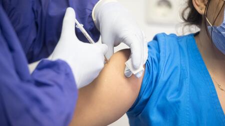 Die mit Handschuhen bekleideten Hände einer Person, die einer Frau den Impfstoff in den Arm spritzt.