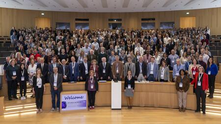 Das Bild zeigt eine große Gruppe von Menschen in einem modernen Hörsaal. Viele der Teilnehmenden tragen Namensschilder an blauen Bändern. Im Vordergrund steht ein Schild mit der Aufschrift „Welcome! DZIF Annual Meeting 2023“.