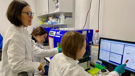 Two female researchers in white lab coats, one sitting at a lab bench and one standing behind her, discuss the results of a cell examination on a computer screen. Sitting next to them, another researcher is working on another piece of laboratory equipment.