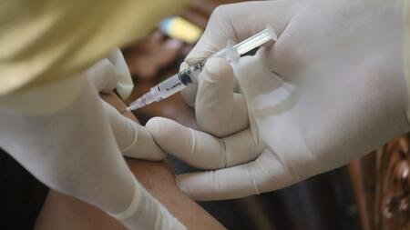 A close-up image of a healthcare professional wearing white gloves administering a vaccine injection into a person's upper arm using a syringe.