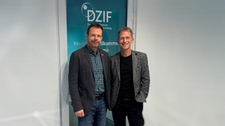 Two men in jackets (Timo Jäger on the left and Holger Zimmermann on the right) in front of a door covered in blue foil, with the DZIF logo visible above their heads.