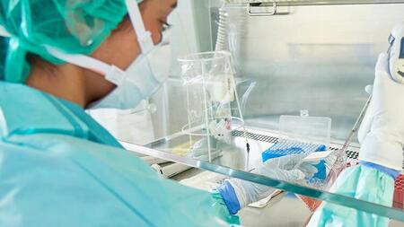 Researcher wearing a cap, mask, and lab coat pipetting at a sterile bench