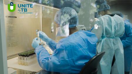 A lab technician at a sterile bench and two others standing in the background, all wearing lab coats, gloves and sterile head covers. The person at the bench is holding a tube in the hand, visible on the bench is a stand with many tubes with black screw caps.