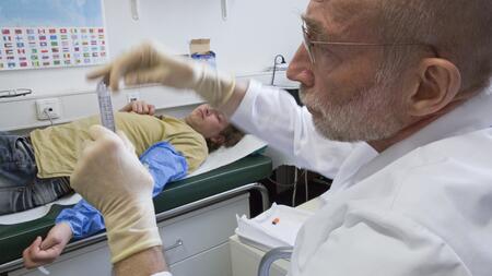 Ein Mann in weißem Kittel und mit medizinischen Handschuhen prüft eine gläserne Spritze mit klarer Flüssigkeit. Im Hintergrund liegt ein männlicher Patient mit einem blauen Überzieher über dem ausgestreckten linken Arm.