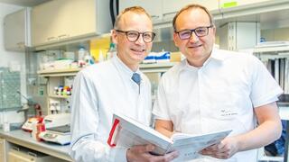 Prof. Wedemeyer und Prof. Cornberg stehen nebeneinander. Sie halten gemeinsam ein Laborbuch in den Händen. Im Hintergrund ist, leicht verschwommen, ein Labor zu erkennen.