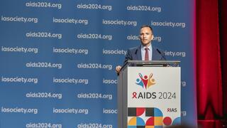 Prof. Christoph Spinner stands at a lectern in a dark blue suit with a red tie and speaks into a microphone. The lectern bears the logo and the words "AIDS 2024". A blue wall can be seen in the background, on which the texts "aids2024.org" and "iasociety.org" are repeatedly printed. A red curtain is visible on the right of the picture.