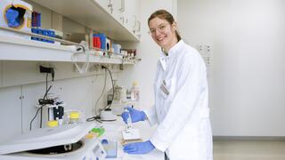 A scientist is working in a laboratory. She is wearing glasses, a white lab coat and blue gloves, and is holding a pipette as she works with samples. In the background are laboratory equipment and shelves of scientific supplies.