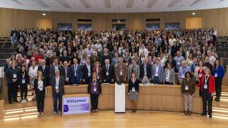 The picture shows a large group of people in a modern lecture hall. Many of the participants are wearing name tags on blue ribbons. In the foreground, there is a sign that reads “Welcome! DZIF Annual Meeting 2023”.
