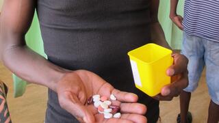 Partial view of a dark-skinned man holding different colored pills in the palm of his right hand and a yellow plastic box in his left hand