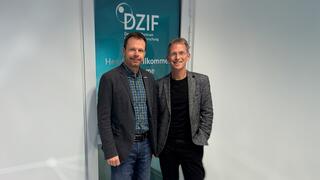 Zwei männliche Personen in Sakkos (links Timo Jäger und rechts Holger Zimmermann) vor einer Tür mit blauer Folie, über ihren Köpfen ist das Logo des DZIF zu erkennen.