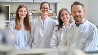 The photo shows a group of four researchers (three women and one man) in white lab coats in a laboratory. 