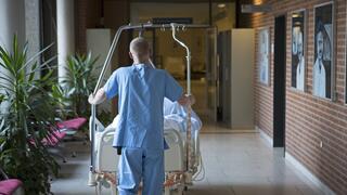 Ein Mann in einem Kasack, mit dem Rücken zum Betrachtenden, schiebt ein Bett mit einem Patienten/einer Patientin durch einen Krankenhausflur. Ebenfalls zu sehen ist ein Wartebereich mit Palmen auf der linken Seite und eine rote Backsteinwand mit Porträts in schwarzweiß auf der rechten Seite. Der Mann schiebt das Bett auf eine geöffnete Glastür zu, hinter der ein beleuchteter Flur liegt.