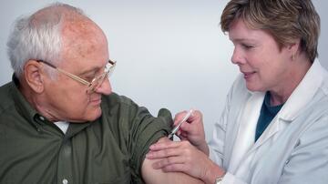 A female doctor wearing a white coat injects the upper arm of an elderly man.