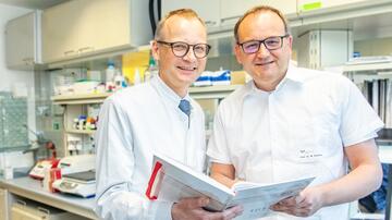 Prof. Wedemeyer und Prof. Cornberg stehen nebeneinander. Sie halten gemeinsam ein Laborbuch in den Händen. Im Hintergrund ist, leicht verschwommen, ein Labor zu erkennen.