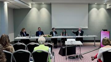 Das Bild zeigt eine Podiumsdiskussion bei einer Konferenz. Fünf Personen sitzen an einem langen Tisch, während eine Person spricht und gestikuliert. Im Vordergrund sitzen einige Zuhörerinnen und Zuhörer. Rechts steht ein Rednerpult mit der Aufschrift „DGI / DZIF Joint Meeting 2025“. Der Raum hat eine schlichte Einrichtung mit einem Projektor, einem großen Bildschirm und Wasserkaraffen auf dem Tisch.