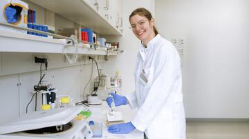 A scientist is working in a laboratory. She is wearing glasses, a white lab coat and blue gloves, and is holding a pipette as she works with samples. In the background are laboratory equipment and shelves of scientific supplies.