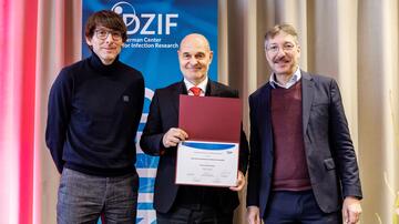 The picture shows the award ceremony at the Joint Annual Meeting of the DZIF and DGI. Three men are standing together, the middle one is holding a certificate with the title “DZIF Prize for Translational Infection Research 2024”. They are all smiling and wearing formal attire. The DZIF logo can be seen on a roll-up in the background.