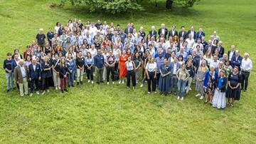 The picture shows a large group of people standing on a green meadow and smiling at the camera. The group consists of men and women of different ages and different clothing. Some are wearing formal clothing such as suits and blouses, while others are dressed more casually.