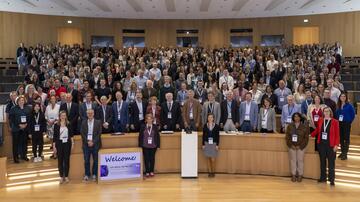 The picture shows a large group of people in a modern lecture hall. Many of the participants are wearing name tags on blue ribbons. In the foreground, there is a sign that reads “Welcome! DZIF Annual Meeting 2023”.