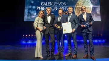 Zwei der vier Gewinner des Galenus-von-Pergamon-Preises in der Kategorie Grundlagenforschung - Dr. Alexander Simonis (Mitte) und Prof. Dr. Florian Klein (2. v. r.) bei der Preisverleihung. Überreicht wurde der Galenus-Preis von Professor Thomas Eschenhagen (2. v. l.). Moderiert wurde die Gala von Yve Fehring (links) und Matthias Gabriel (rechts). Die Personen stehen nebeneinander auf einer Bühne in festlicher Kleidung.