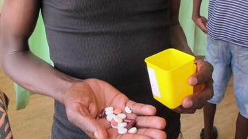 Teilansicht eines dunkelhäutigen Mannes, der verschiedenfarbige Pillen auf der Handfläche seiner rechten Hand hält und in der linken Hand eine gelbe Plastikdose 