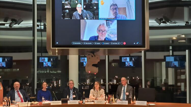 Mitglieder des PKAMR in einem Sitzungssaal, andere Personen sind über einen großen Monitor zugeschaltet