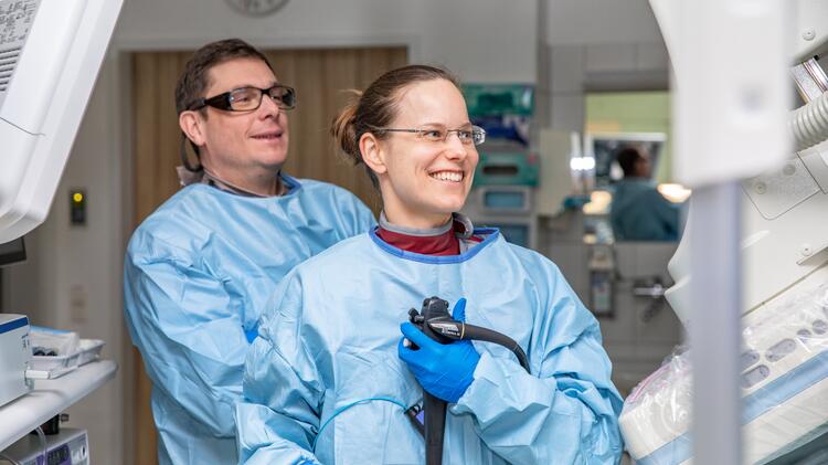 A man and a woman in blue surgical gowns and blue gloves are looking at a monitor. The woman is holding medical equipment in her hands. They are surrounded by other medical equipment.