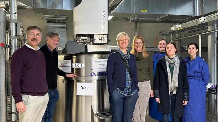 Seven people standing in a laboratory room next to a large metal device, with pipes, ducts, and technical equipment visible in the background.