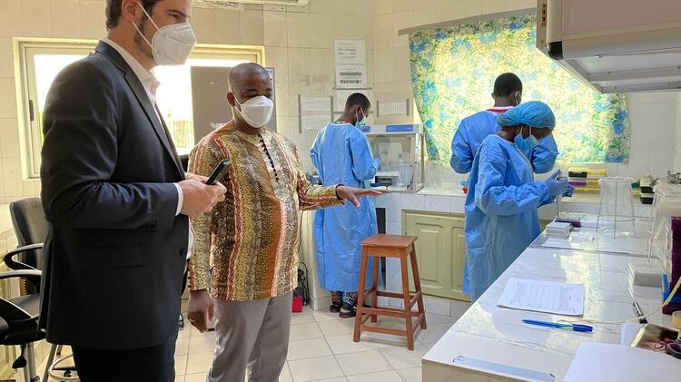 Laboratory of Viral Hemorrhagic Fever (LFHB) in Benin. Prof. Jan Felix Drexler and Dr. Anges Yadouleton are standing on the left, half facing the viewer. In the background, three lab workers wearing blue lab coats and gloves (with one of them additionally wearing a blue head cover) working at lab benches.