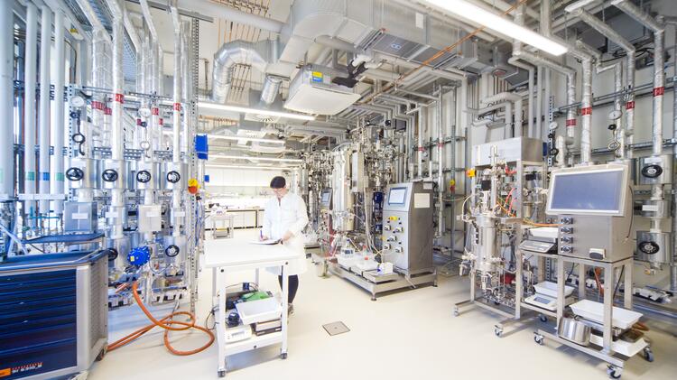 Forschungsumgebung mit Fermentationsanlagen: Eine Wissenschaftlerin in Laborkleidung arbeitet an einem Tisch in einem technisch ausgestatteten Labor mit Edelstahlreaktoren, Rohrleitungen und Steuerungssystemen.