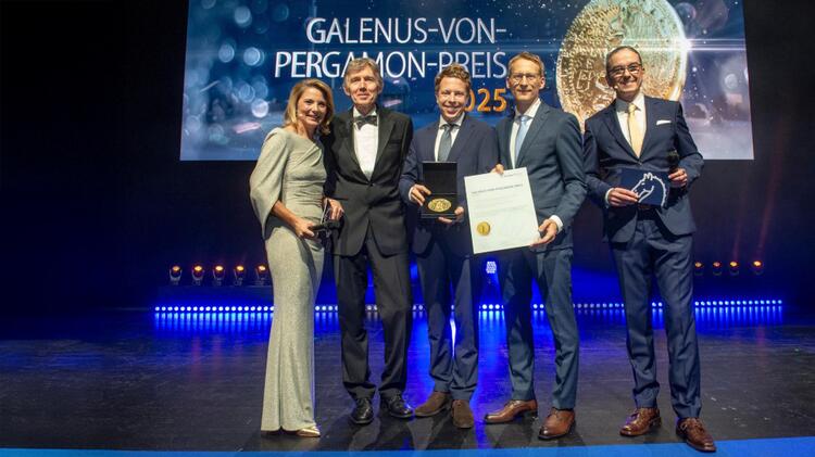 Zwei der vier Gewinner des Galenus-von-Pergamon-Preises in der Kategorie Grundlagenforschung - Dr. Alexander Simonis (Mitte) und Prof. Dr. Florian Klein (2. v. r.) bei der Preisverleihung. Überreicht wurde der Galenus-Preis von Professor Thomas Eschenhagen (2. v. l.). Moderiert wurde die Gala von Yve Fehring (links) und Matthias Gabriel (rechts). Die Personen stehen nebeneinander auf einer Bühne in festlicher Kleidung.
