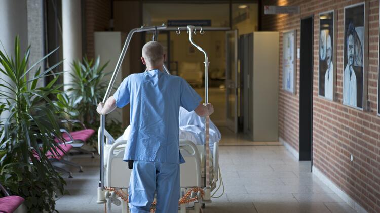 A man in a smock, with his back to the viewer, pushes a bed with a patient through a hospital corridor. Also visible is a waiting area with palm trees on the left and a red brick wall with black-and-white portraits. The man pushes the bed toward an open glass door, behind which is an illuminated corridor.